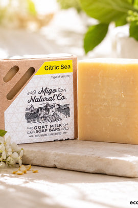 A bar of Citric Sea goat milk soap by Migo Natural Co. stands next to its kraft paper packaging box on a light stone surface, with soft-focus greenery and white flowers in the background.