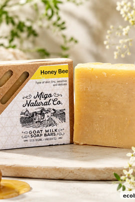 A bar of Migo Natural Co. Honey Bee Goat Milk Soap is displayed next to its kraft paper packaging box on a wooden board, with a honey dipper and soft floral background props.