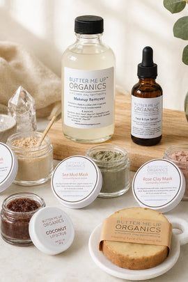 A curated flat-lay of Butter Me Up Organics skincare products on a light stone surface with a wooden board and soft natural styling. Visible items include a large clear bottle of Makeup Remover, a small amber dropper bottle, several round jars of masks and scrubs, a wrapped coconut body scrub, and a wrapped bar of soap, arranged with fabric, crystals, and greenery.