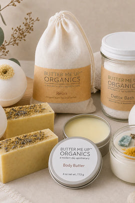 A curated flat-lay of Butter Me Up Organics bath and body products on a neutral fabric surface, including a drawstring bag labeled Relax, a jar labeled Detox Bath, a tin labeled Body Butter, a small candle jar, and two bath bombs with dried flower centers.