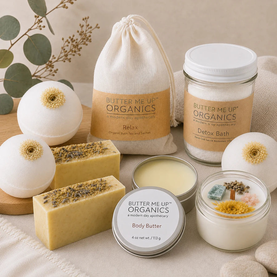 A curated flat-lay of Butter Me Up Organics bath and body products on a neutral fabric surface, including a drawstring bag labeled Relax, a jar labeled Detox Bath, a tin labeled Body Butter, a small candle jar, and two bath bombs with dried flower centers.