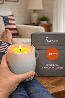 Person holding a lit candle next to a 'Sanari' biscotti candle box in a cozy living room.
