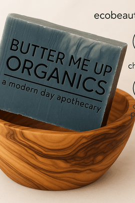  soap bar labeled 'Butter Me Up Organics' displayed in a carved wooden bowl with the text ecobeaut.com in the top right corner and icons reading 'no chemicals' and 'organic products' on a neutral background.